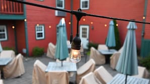 An outdoor seating area with tables and chairs covered in beige cloths, connected by a string of lights.