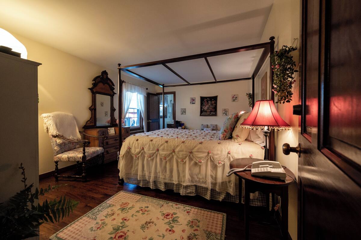 A four-poster bed with a floral bedspread, a bedside lamp, a chair, and a small table with a telephone.