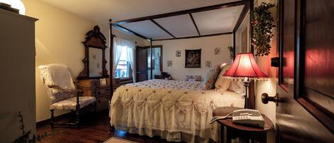 A four-poster bed with a floral bedspread, a bedside lamp, a chair, and a small table with a telephone.