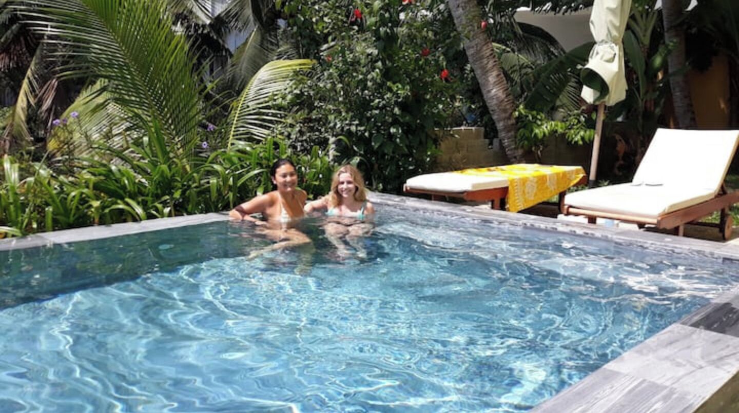 A swimming pool with two people in the water, surrounded by lush greenery and a sunbed.