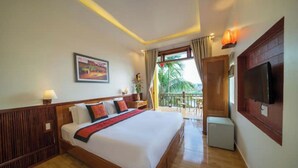 A hotel room with a large bed, a TV, a balcony with a view of palm trees, and a painting on the wall. - Deluxe Double Room with Balcony, Lake, River View (Hoi An)