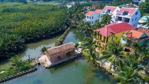 A coastal property with a thatched-roof structure and colorful buildings, surrounded by water and palm trees. - Deluxe Double Room with Balcony, Lake, River View (Hoi An)