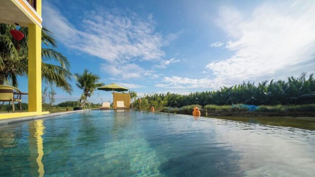A clear pool with a yellow building on one side, palm trees, and a view of the sky. - Deluxe Double Room with Balcony, Lake, River View (Hoi An)