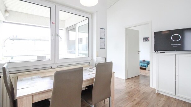 A modern, well-lit room with a large window, a desk with chairs, and a television on a cabinet. - nJoy! Sunny Apartment Göppingen | Balcony & Parking (Göppingen)