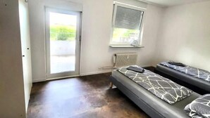 Ein kleiner, moderner Raum mit einem Bett, einem Fenster mit Jalousien, einer Heizung und einer Tür zu einem Außenbereich.