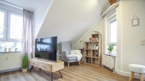 Una sala de estar moderna con un televisor sobre un mueble de madera, un sillón gris, una butaca blanca y mullida, y una estantería.