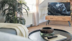 Una sala de estar con un televisor sobre un mueble de madera, una mesa de centro con un control remoto, una planta y una estantería con libros y una lámpara.