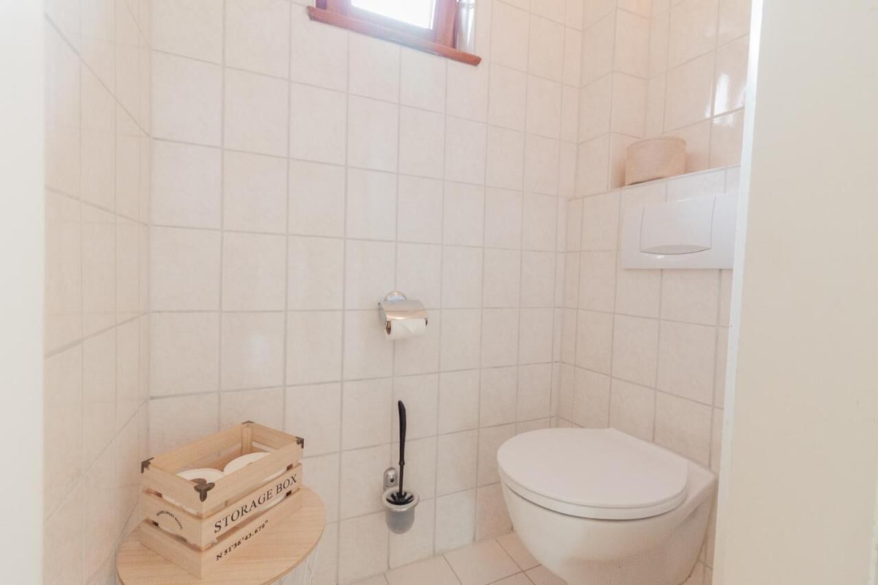 Ein kleines, sauberes Badezimmer mit weißen Fliesen, einer Toilette, einer Aufbewahrungsbox und einem Pömpel.