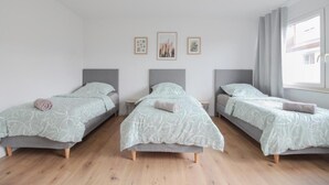 Ein Zimmer mit drei Einzelbetten, jedes mit grauem Kopfteil und einer hellgrünen, gemusterten Bettdecke. Darüber hängen drei gerahmte Bilder an der Wand.