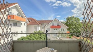 Ein Balkon mit zwei Holzstühlen und einem kleinen Tisch, mit Blick auf benachbarte Gebäude und Grünflächen.