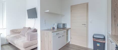 A modern, minimalistic room with a wooden floor, a beige sofa, a small kitchenette with a microwave, and a TV mounted on the wall.