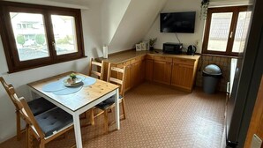 Een kleine keuken met een eettafel, stoelen, een aan de muur bevestigde televisie, en een raam dat voor natuurlijk licht zorgt.