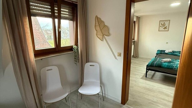 Een kamer met twee witte stoelen, een raam met zonwering, een decoratief blad aan de muur en een bed in de aangrenzende kamer.