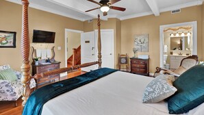 A bedroom with a four-poster bed, a ceiling fan, a television, a dresser, and a vanity with a mirror.