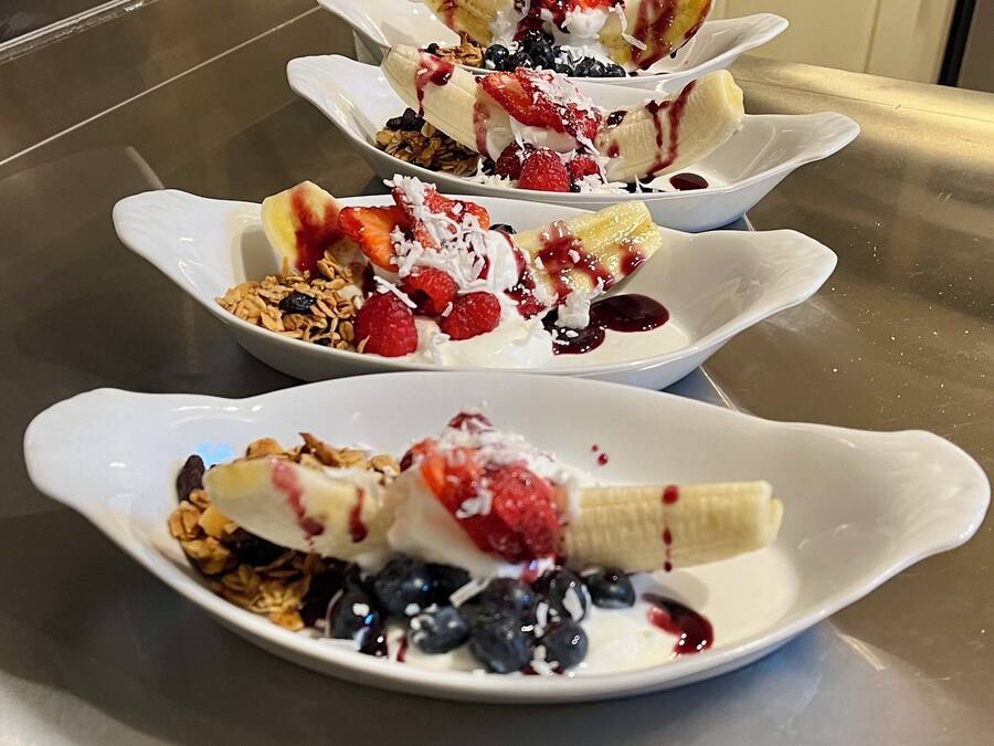 Tres tazones blancos con un postre de plátanos, bayas y granola sobre una superficie de acero inoxidable.