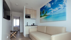 A modern living room with a beige sofa, a kitchen with a microwave, and a large tropical beach painting on the wall. - Magic Pirates Island Resort (Benidorm)