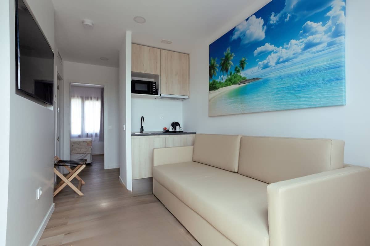 Una sala de estar moderna con un sofá color beige, una cocina con microondas y un cuadro grande de una playa tropical en la pared.