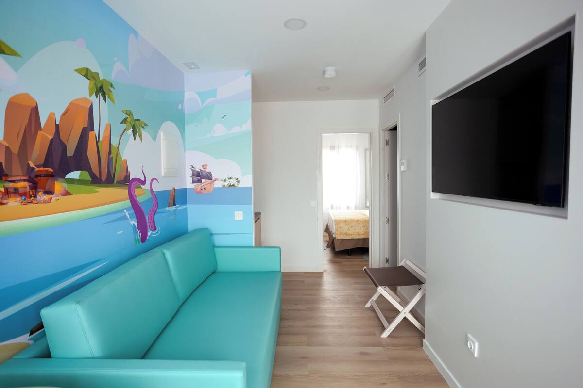 Una habitación con un sofá color turquesa, un televisor grande y un mural que representa una escena de playa.