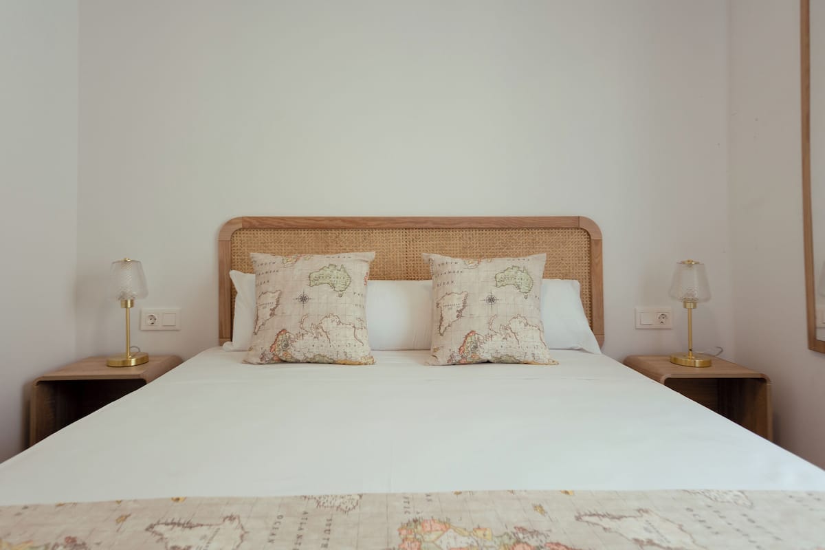 Una cama bien tendida con sábana blanca y dos almohadas decorativas con estampado de mapa, flanqueada por dos mesitas de noche con lámparas.