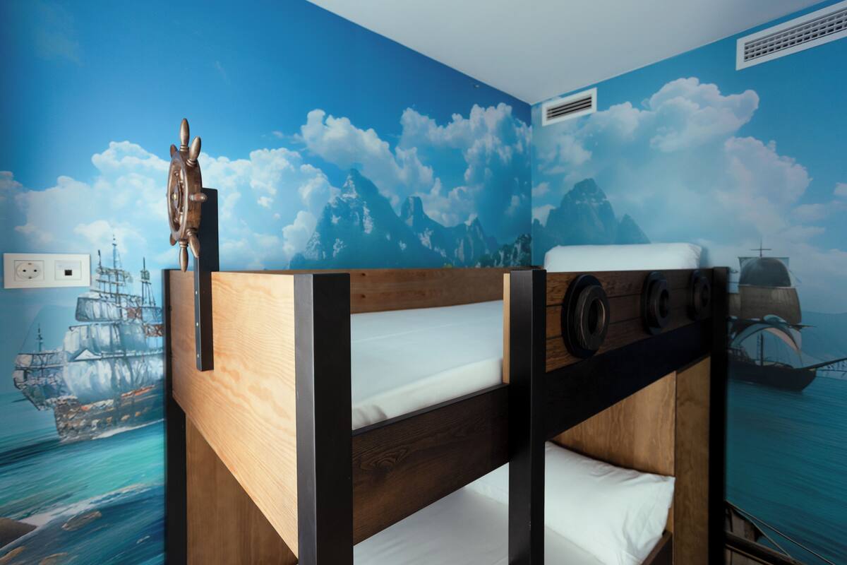 Una cama nido con estructura de madera y ropa de cama blanca, frente a una pared con un mural marítimo que muestra barcos y montañas.