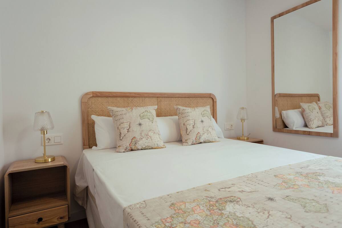 Una cama bien hecha con una colcha blanca y dos almohadones decorativos, una mesita de noche de madera con una lámpara y un espejo grande en la pared.