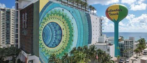 Um hotel com um grande mural na fachada, vista para o mar e uma placa indicando 'Hollandale Beach'.