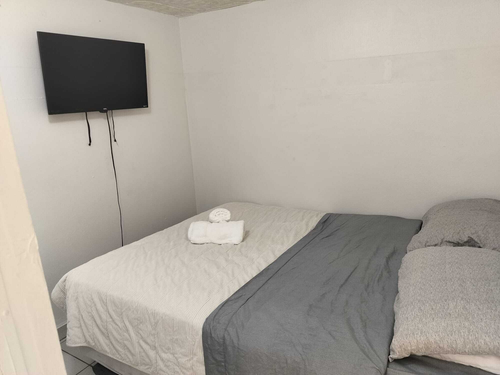 Una cama individual con edredón blanco y gris, una toalla blanca enrollada y una almohada gris. Un televisor de pantalla plana está montado en la pared sobre la cama.