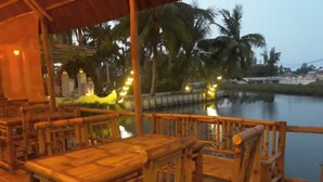 A wooden deck with tables and chairs overlooking a calm body of water, surrounded by palm trees and a fence.