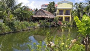 A tropical setting with a pond, a thatched-roof hut, and a yellow building with balconies.