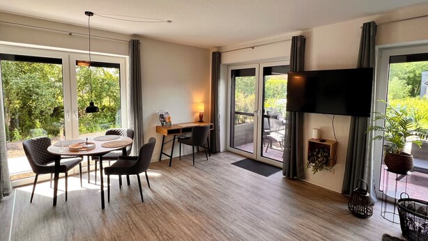 A modern room with a dining area, a desk with a lamp, a TV, and large windows offering a view of greenery.