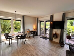 A modern room with a dining area, a desk with a lamp, a TV, and large windows offering a view of greenery. - New comfort apartment Nordic at Nature, Emden by Fewo & Zimmer Bruns (Emden)