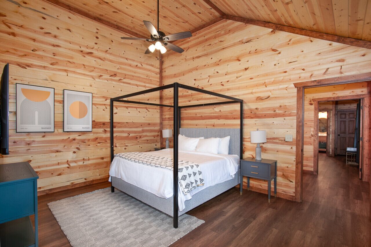 A four-poster bed in a log cabin-style room with wooden walls, a ceiling fan, and a small bedside table with a lamp.