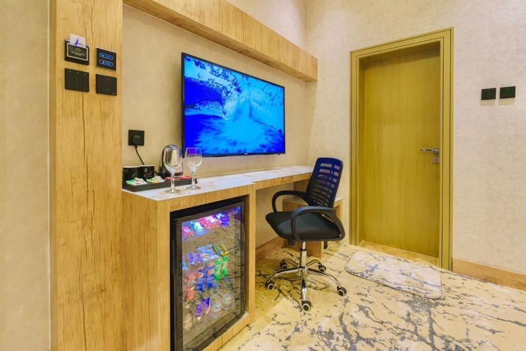 Una habitación de hotel con un escritorio de madera, un televisor de pantalla plana, un aparador de vidrio con objetos decorativos y una puerta.