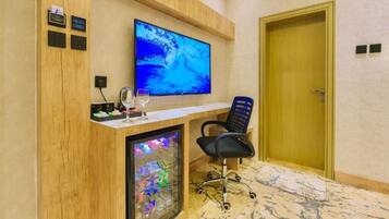 Una habitación de hotel con un escritorio de madera, un televisor de pantalla plana, un aparador de vidrio con objetos decorativos y una puerta.