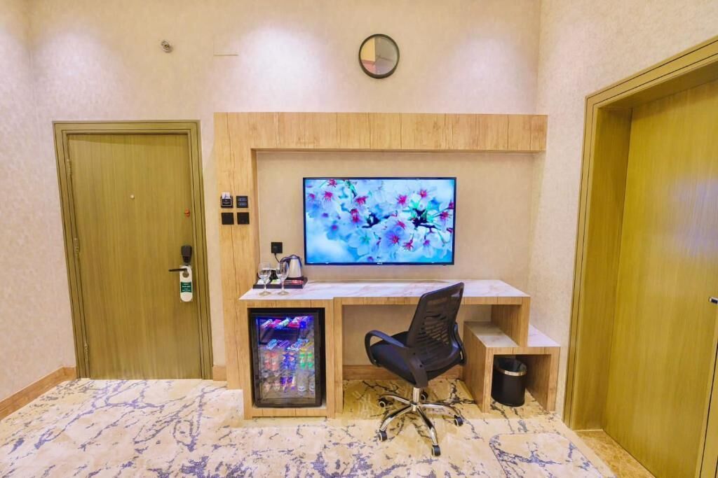 Hotellihuoneessa on puinen työpöytä, tietokonenäyttö, tuoli, pieni jääkaappi ja seinällä oleva kello.