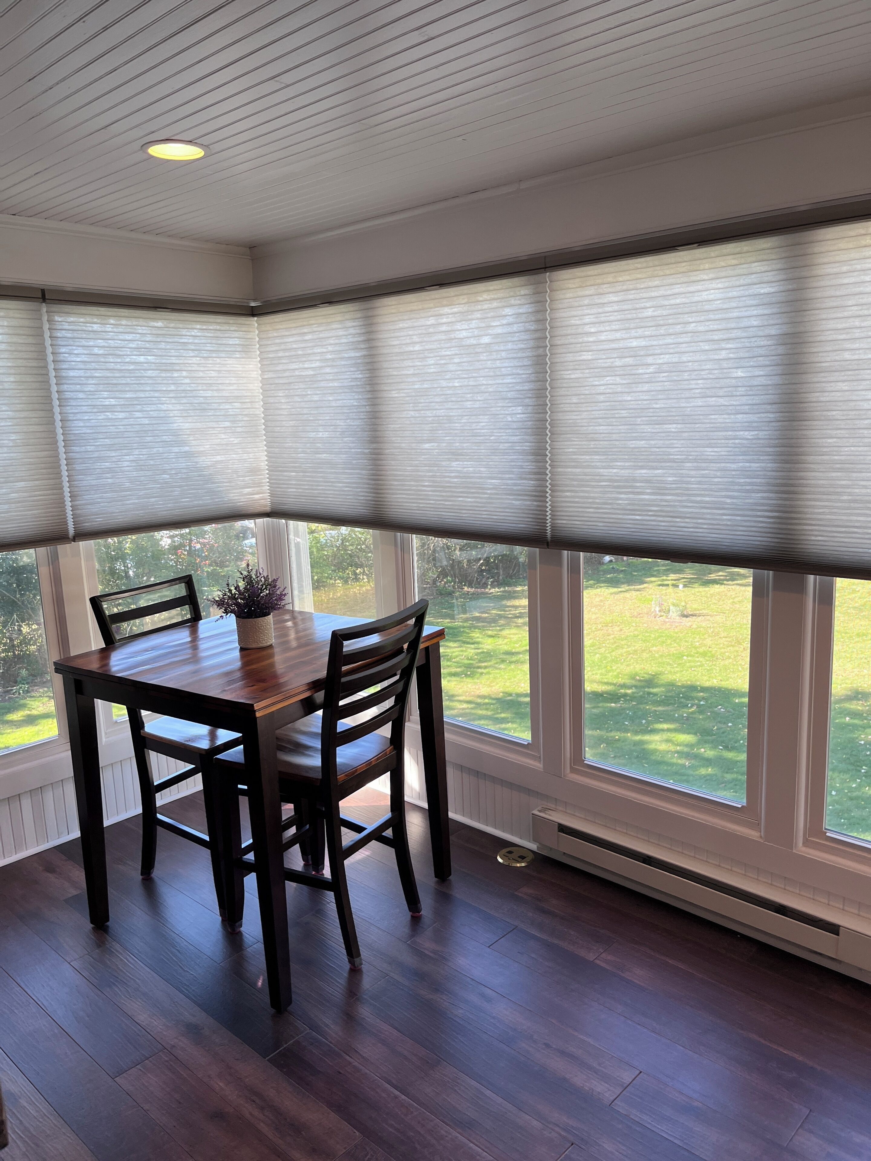 A dining area with a wooden table and chairs, large windows with blinds, and a view of a green landscape.