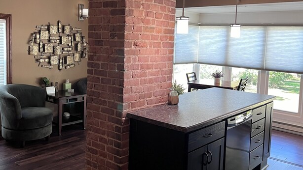 Une cuisine moderne avec un mur d’accent en briques, un îlot central en bois foncé doté de tiroirs, et un coin repas avec une chaise et une table.