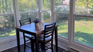 Un espace repas avec une table et des chaises en bois, de grandes fenêtres donnant sur une pelouse verdoyante et des arbres, et un plafond doté d’un éclairage encastré.
