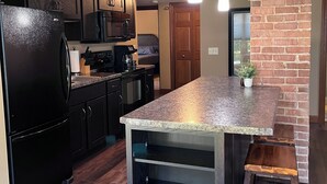 Une cuisine moderne avec des meubles de couleur sombre, un réfrigérateur noir, un plan de travail en granit et un mur d’accent en briques.