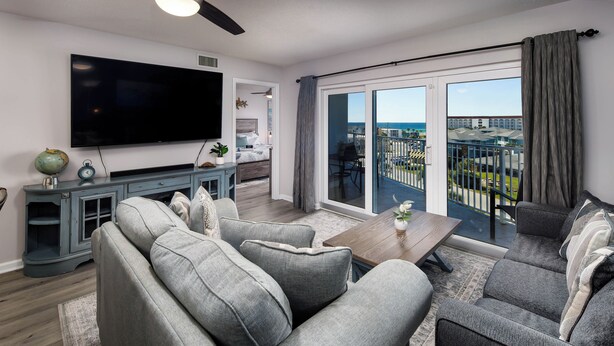 A modern living room with a large flat-screen TV, grey sofas, a wooden coffee table, and a balcony with a view of the ocean. - Gulf View Island 2/2 Condo w/ Restaurant Nearby (Fort Walton Beach)