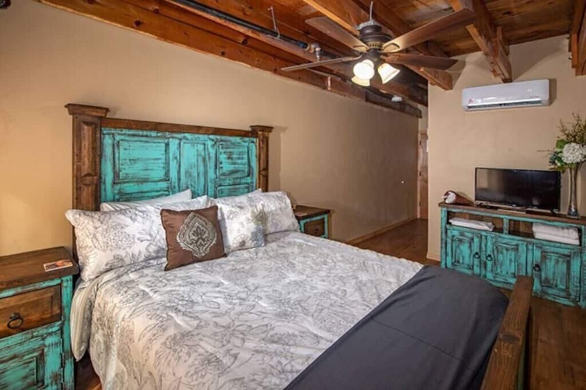 A bedroom with a wooden bed frame, a ceiling fan, an air conditioning unit, a television, and a small nightstand with a lamp.