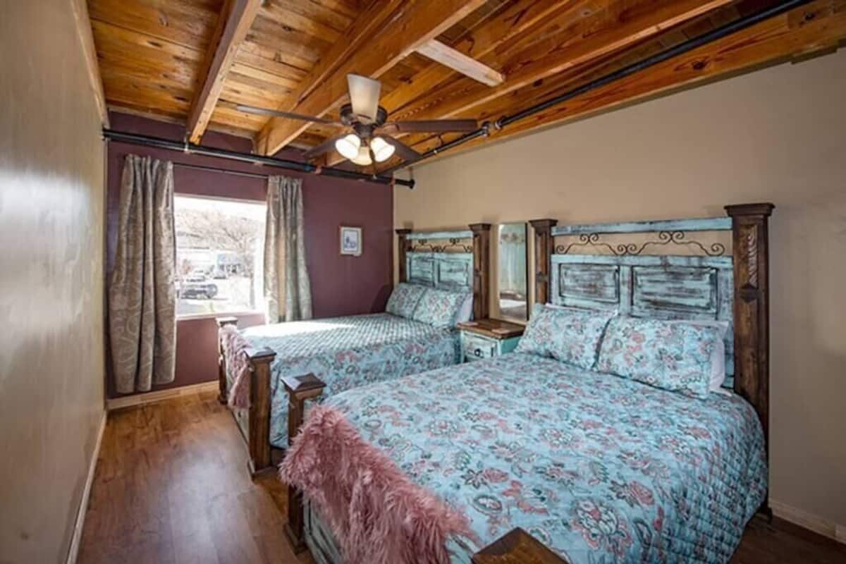 A bedroom with two beds, wooden ceiling beams, a ceiling fan, and a window with curtains.