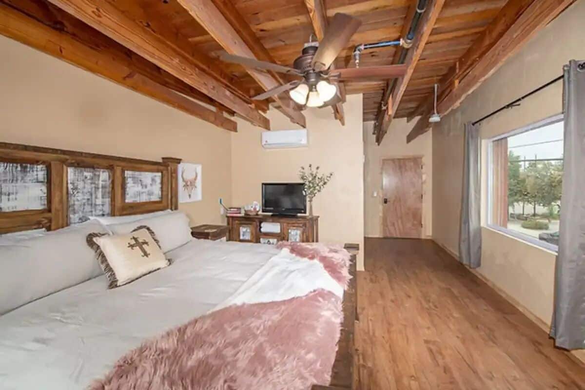 A bedroom with a large bed, wooden ceiling beams, a ceiling fan, a television, and a window with curtains.