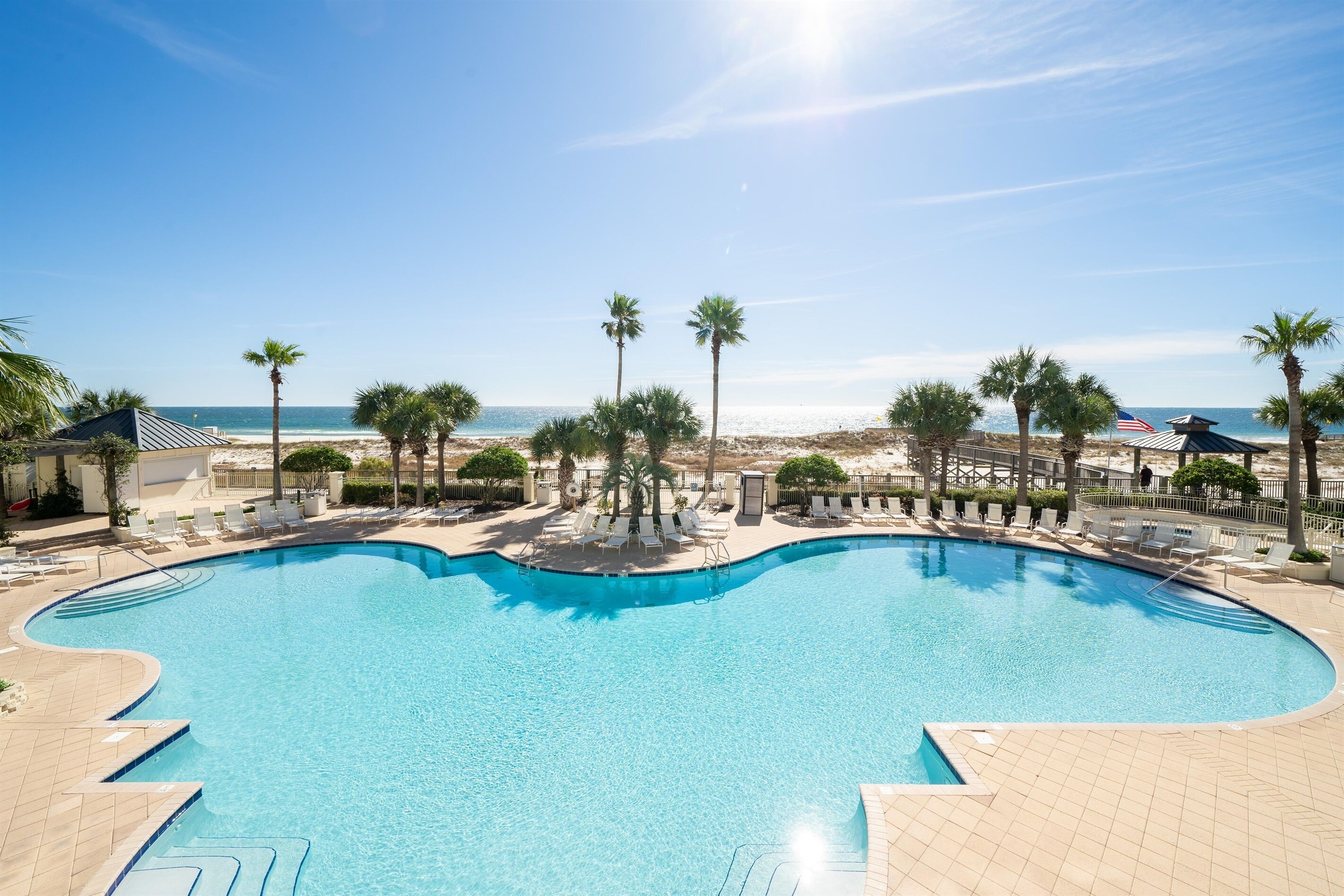 A large, curved swimming pool with clear blue water, surrounded by palm trees and lounge chairs, with a beach and ocean view in the background.