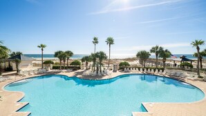 A large, curved swimming pool with clear blue water, surrounded by palm trees and lounge chairs, with a beach and ocean view in the background.