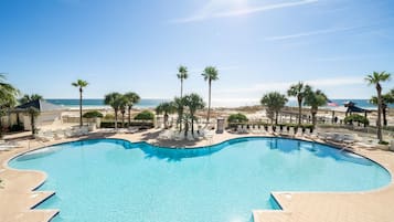 A large, curved swimming pool with clear blue water, surrounded by palm trees and lounge chairs, with a beach and ocean view in the background.