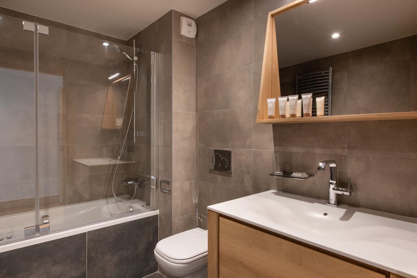 A modern bathroom with a glass-enclosed shower, a toilet, a sink with a mirror, and toiletries on a shelf.