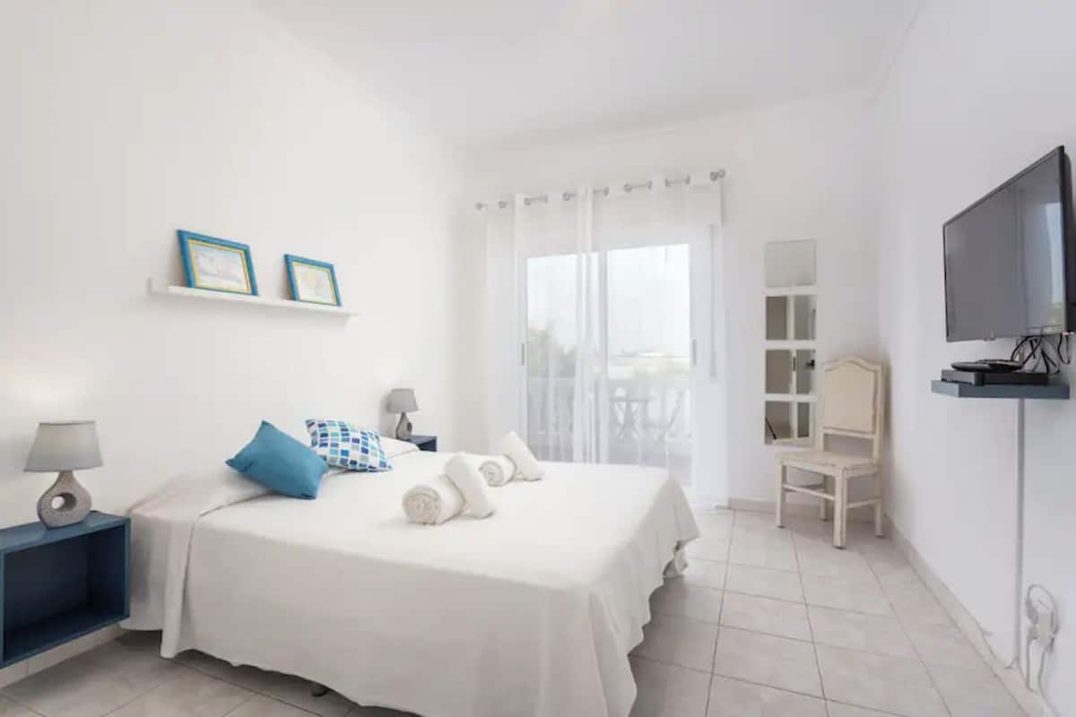 A hotel room with a bed, a TV, a chair, and a balcony with curtains.