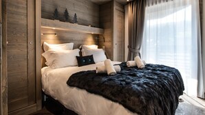 Een moderne hotelkamer met een houten hoofdeinde, een groot bed met witte lakens, een zwarte bont plaid, en een stadszicht door dunne gordijnen.