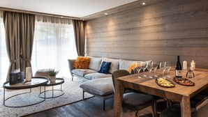 Een moderne woonkamer met een houten wand, een grijze bank met oranje en blauwe kussens, een ronde salontafel en een eettafel gedekt met wijnglazen en een fles.